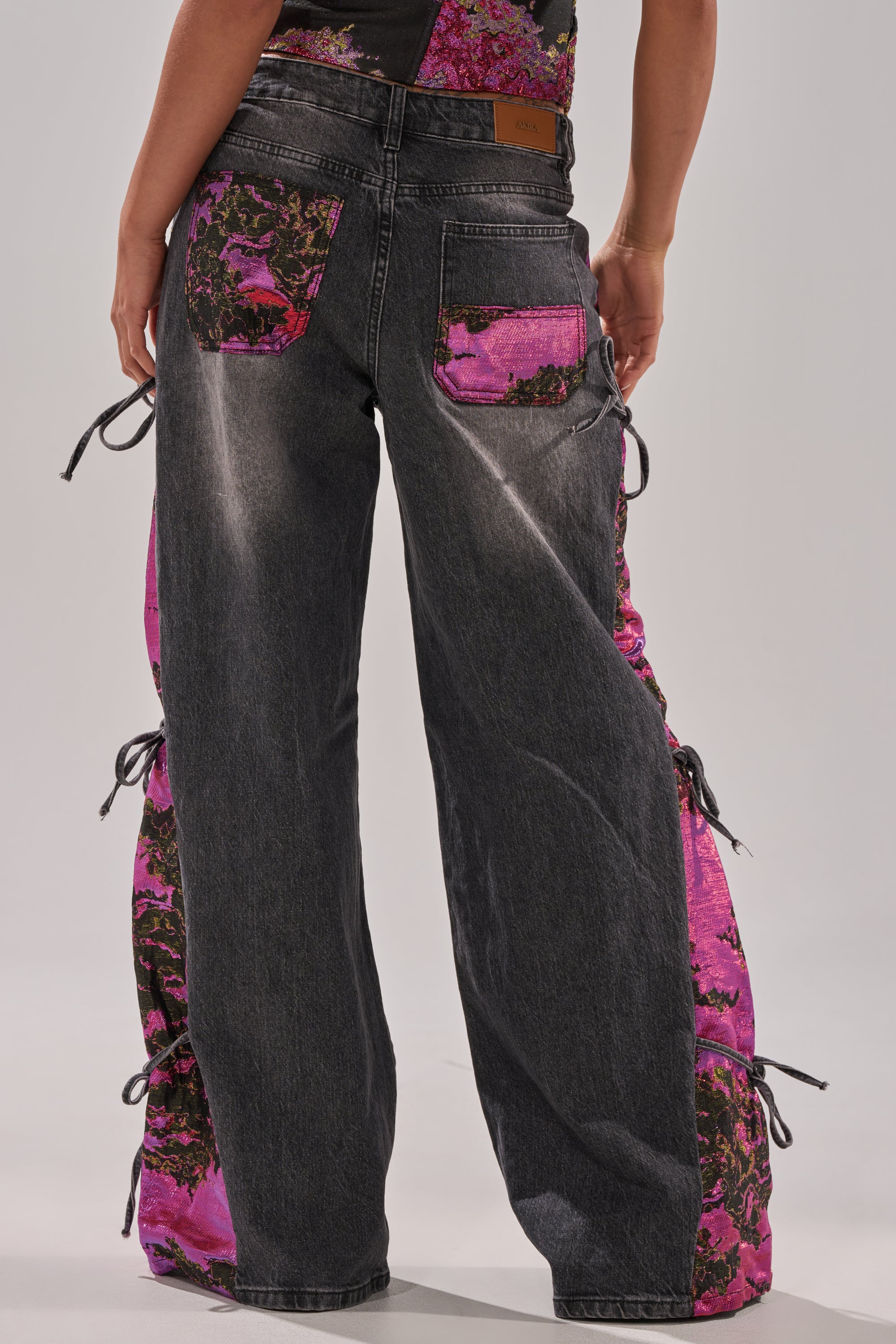 Someone stands with their back to the camera wearing LETS PARTY BROCADE RELAXED DENIM: black wide-leg jeans featuring faded details and bright pink floral patches on the back pockets and sides.