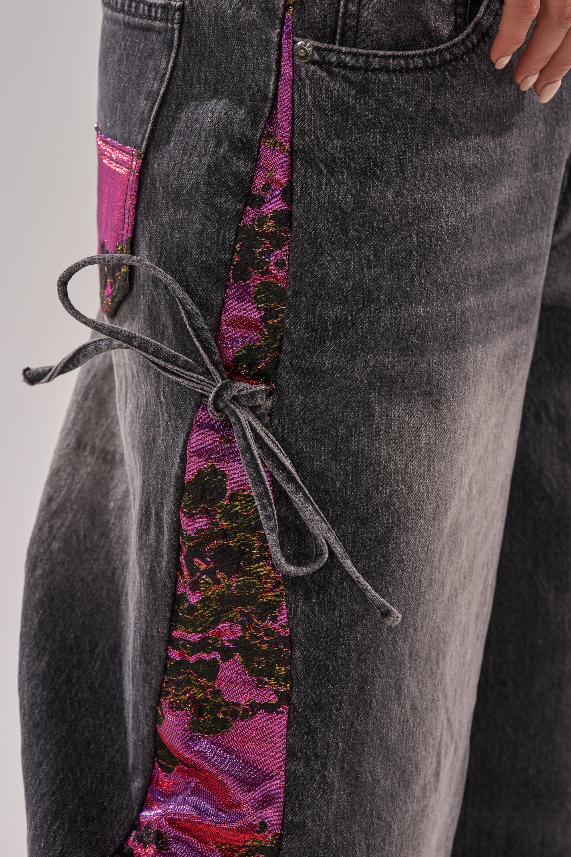 Close-up of LETS PARTY BROCADE RELAXED DENIM: faded black jeans with shiny pink and black floral side panels, metallic patches, gray lace-up detail on the leg, and a manicured hand resting near the pocket.