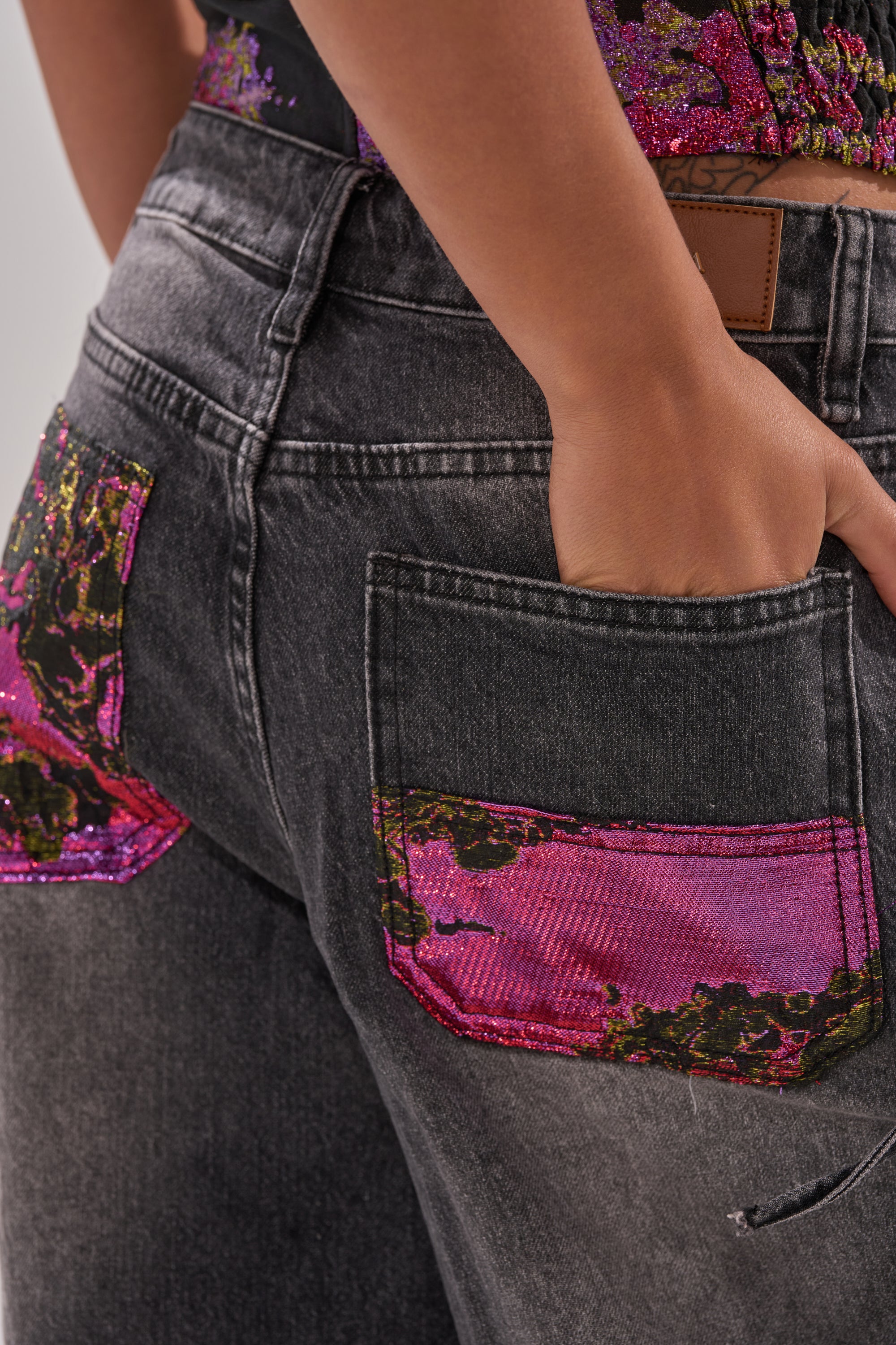 Close-up of a person wearing LETS PARTY BROCADE RELAXED DENIM, featuring wide-leg dark gray jeans with bright pink and green floral sequin details on the back pockets and upper section for a decorative touch.