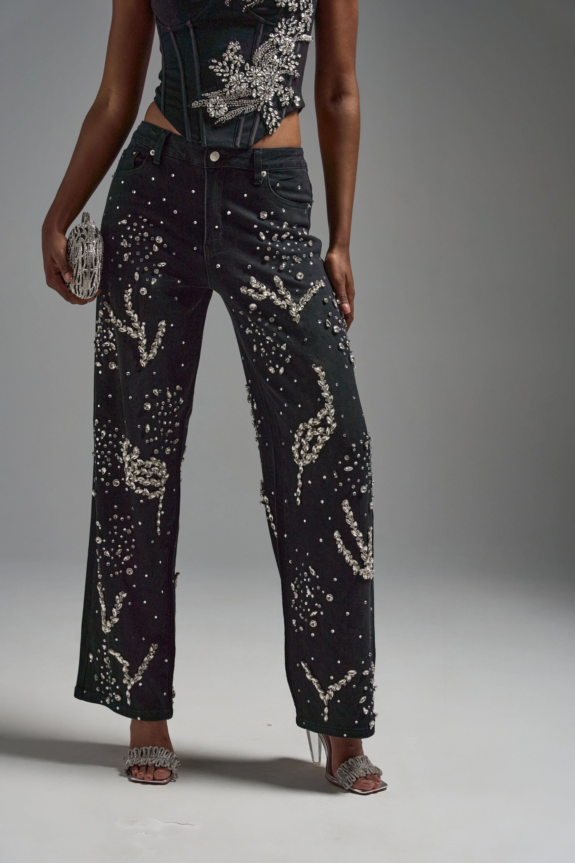 Wearing the GIVE IT TO ME BABY EMBELLISHED DENIM—black rhinestone jeans with sparkling bead patterns—plus a matching embellished corset top, silver heeled sandals, and a silver clutch, standing against a gray background.