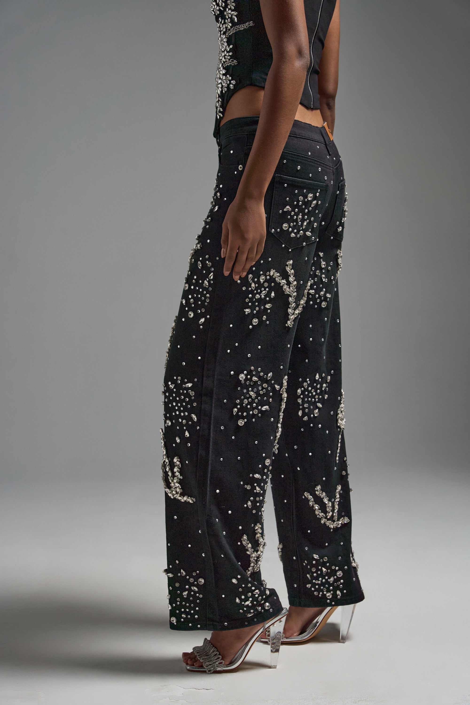 A person stands sideways against a plain gray background wearing the GIVE IT TO ME BABY EMBELLISHED DENIM, high-rise black jeans adorned with rhinestones and pearls, paired with a sleeveless black top and silver high-heeled sandals.