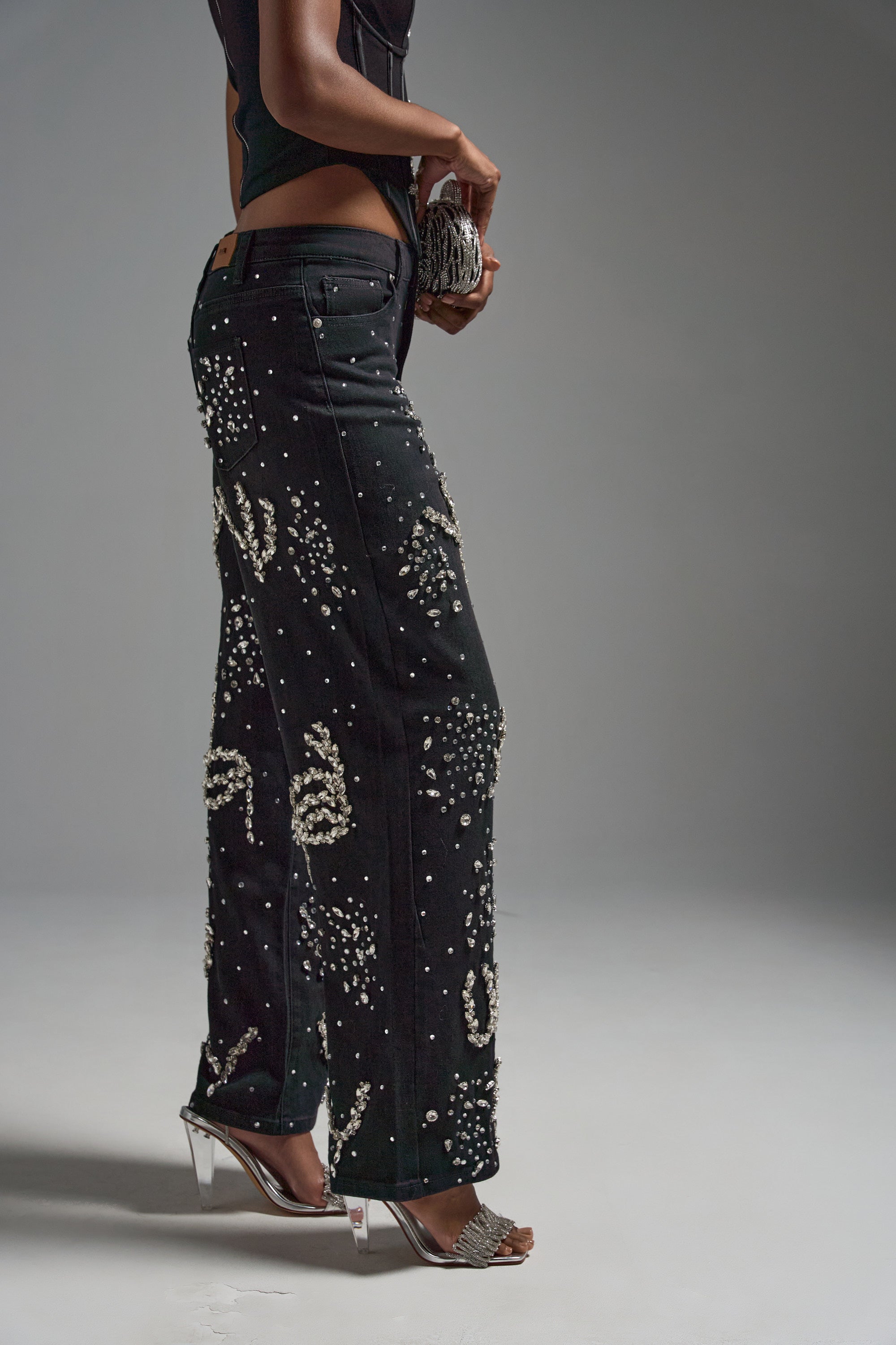 A person wears the GIVE IT TO ME BABY EMBELLISHED DENIM, a black sleeveless top, and silver high-heeled sandals while holding a small silver clutch against a plain gray background.