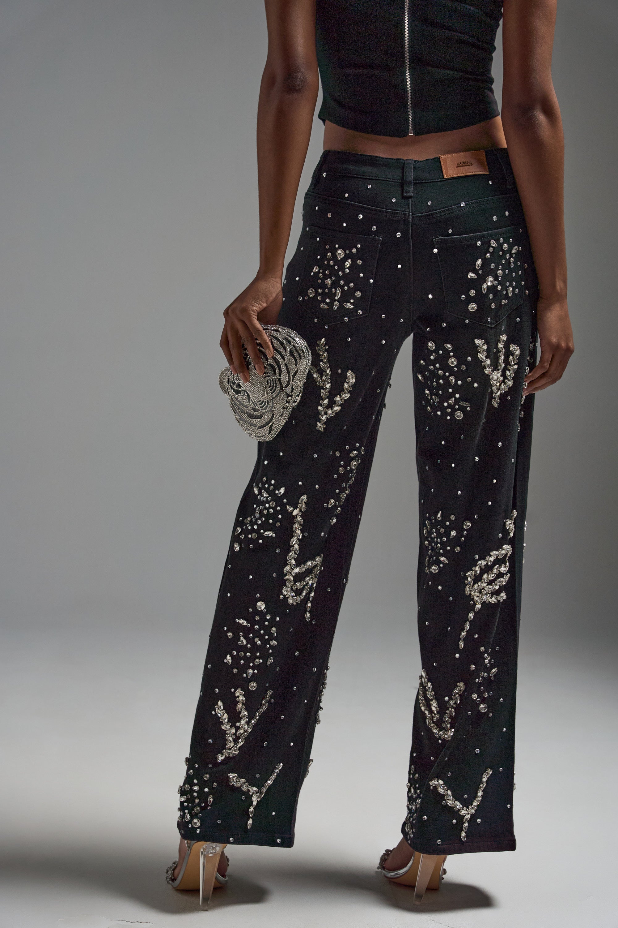 A person faces away from the camera, showcasing the GIVE IT TO ME BABY EMBELLISHED DENIM—high-rise black jeans adorned with pearls and beads—styled with a black sleeveless top, white heels, and a silver clutch against a plain gray background.