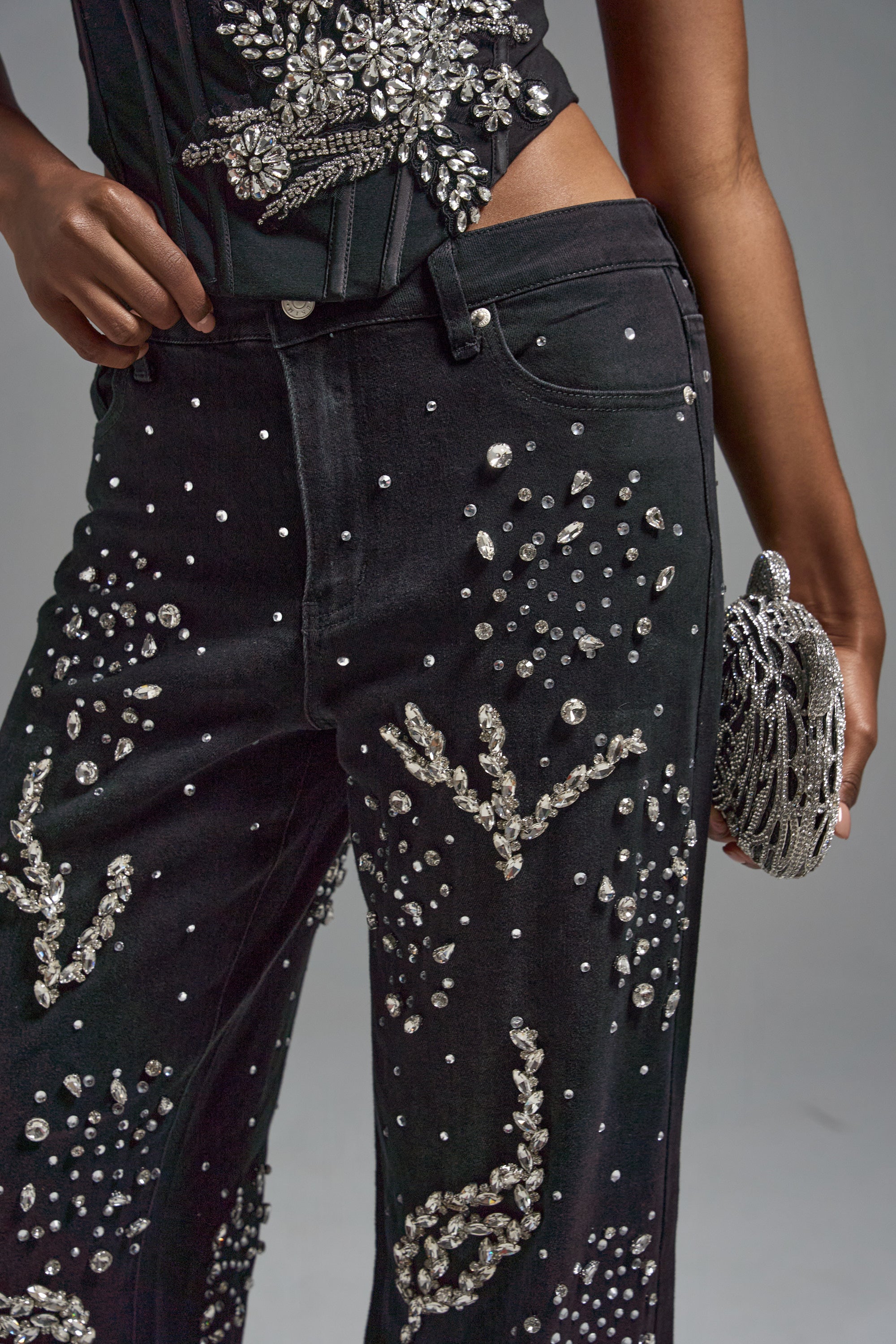 A cropped photo shows a person in GIVE IT TO ME BABY EMBELLISHED DENIM—high-rise black jeans and a matching top with silver beads and rhinestones—holding a small silver beaded clutch, featuring only their lower torso and one arm.