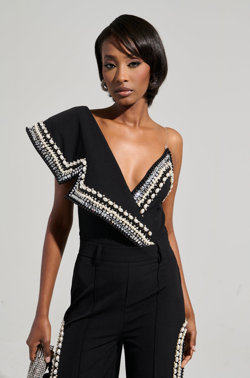 Modeling the Luxurious Asymmetrical Pearl Bodysuit, a person exudes confidence in their fashionable black ensemble adorned with pearls and rhinestones. The look features an asymmetric neckline and sleeve, complemented by their short hairstyle and accessorized with earrings and a matching clutch against a neutral backdrop.