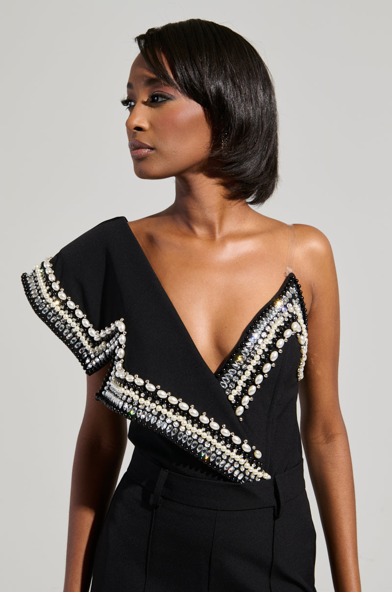 A woman poses against a neutral background wearing the Luxurious Asymmetrical Pearl Bodysuit, which boasts an asymmetrical sleeve and is decorated with pearls, beads, and rhinestones. She gazes to the side with a confident expression, exuding elegance.