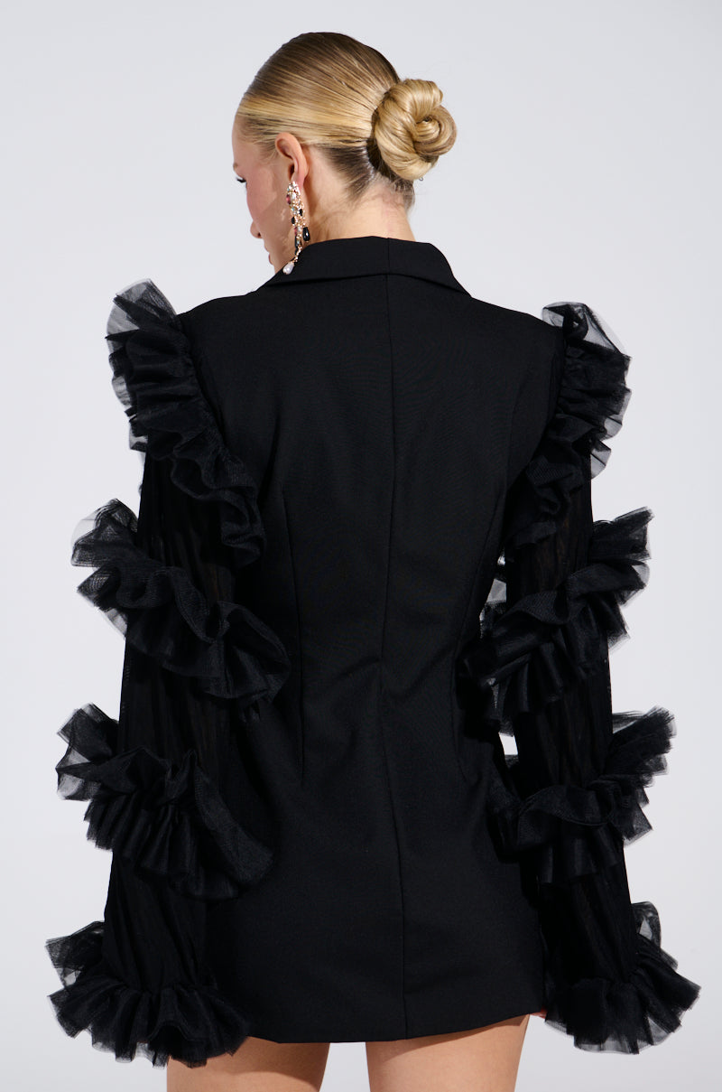 A person with blonde hair in a neat bun exudes luxury fashion, elegantly wearing the LIVING OVER THE TOP RUFFLE SLEEVE BLAZER MINI DRESS. The black mini dress features sheer, ruffled sleeve details as they face away from the camera against a plain background.