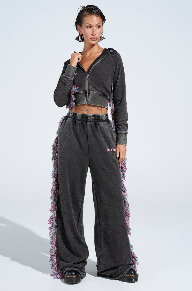 A person in a textured, dark tracksuit with colorful side fringe is donning the midriff-baring zip hoodie and matching wide-leg pants from The One And Only Jogger set. They stand on a light background, wearing black boots.