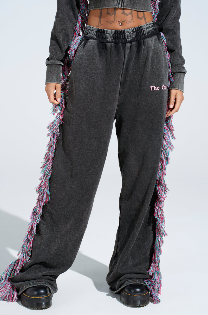 A person wearing "THE ONE AND ONLY JOGGER" in dark gray, featuring colorful fringe detailing and "The Ch" embroidery on the left leg, pairs them with a matching top and chunky black shoes to showcase their midriff.