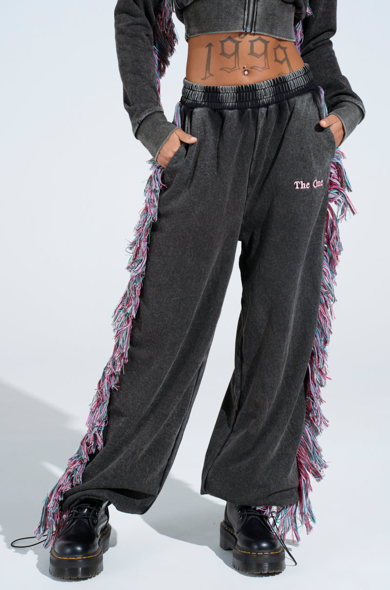 A person confidently poses in grey joggers with colorful fringe detailing, featuring "The One" embroidery on the cotton-blend. Platform shoes and tattoos add flair, bringing "THE ONE AND ONLY JOGGER" to life.