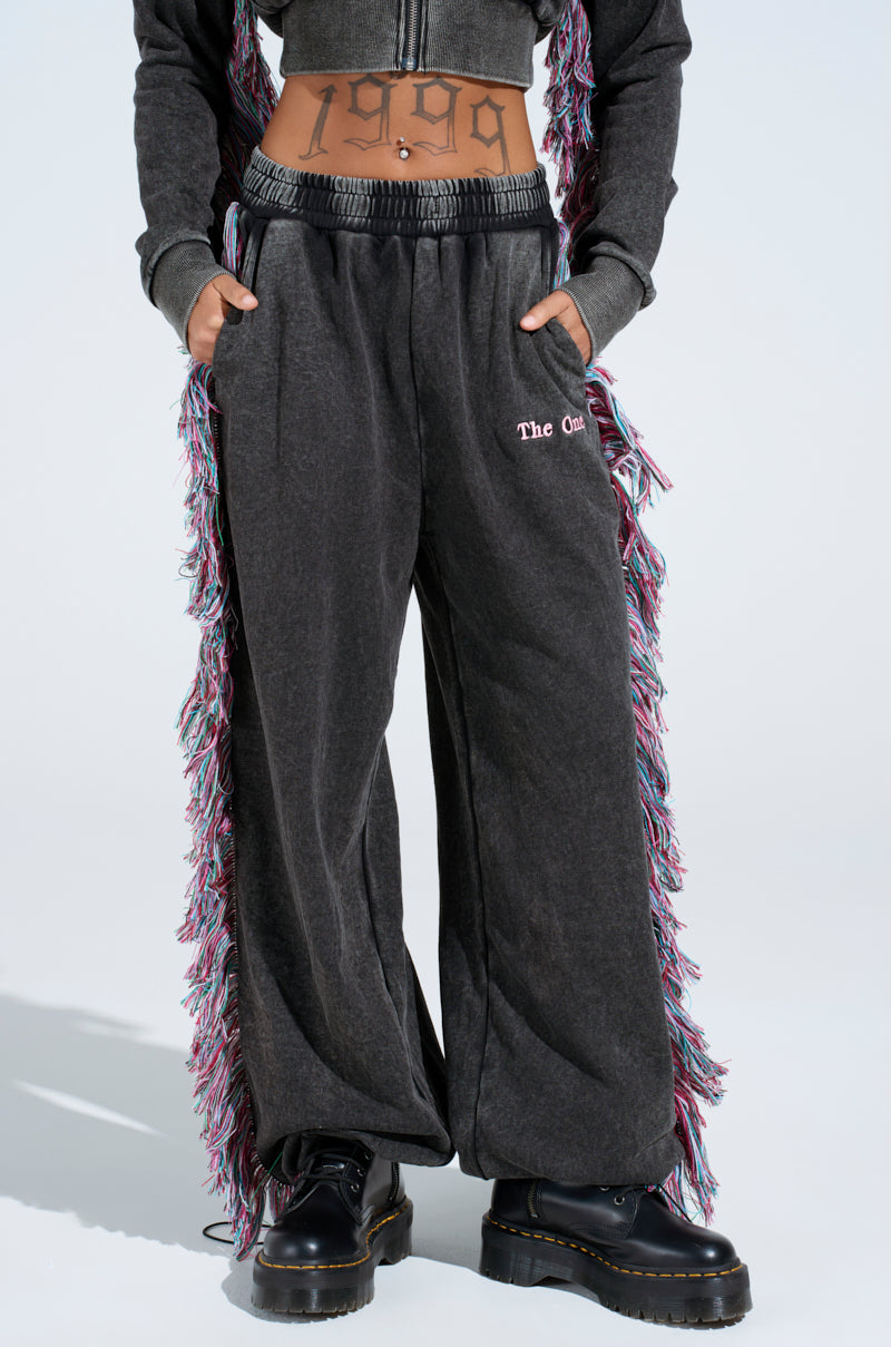 Someone wearing THE ONE AND ONLY JOGGER in dark gray with a pink embroidered pocket logo, a matching crop top, black boots, and outfit adorned with colorful fringe. The year "1999" is tattooed on their stomach as they stand against a light background.