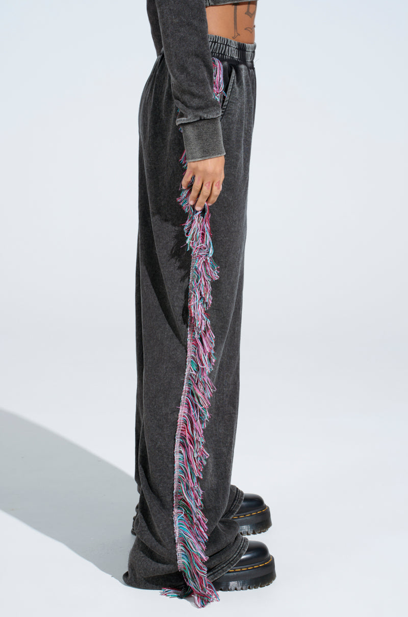 A person models THE ONE AND ONLY JOGGER with colorful fringe down the side, resting their hand on the vibrant trim. Paired with chunky black shoes and set against a crisp white backdrop, these cotton-blend joggers showcase a unique style.
