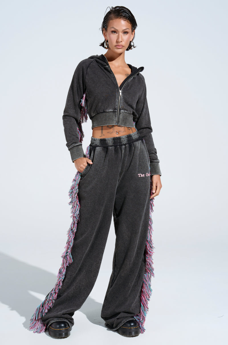 A person confidently wears THE ONE AND ONLY JOGGER, a dark cropped zip-up hoodie with colorful side fringe. This cotton-blend jogger exudes casual style, paired with black shoes against a plain white backdrop.