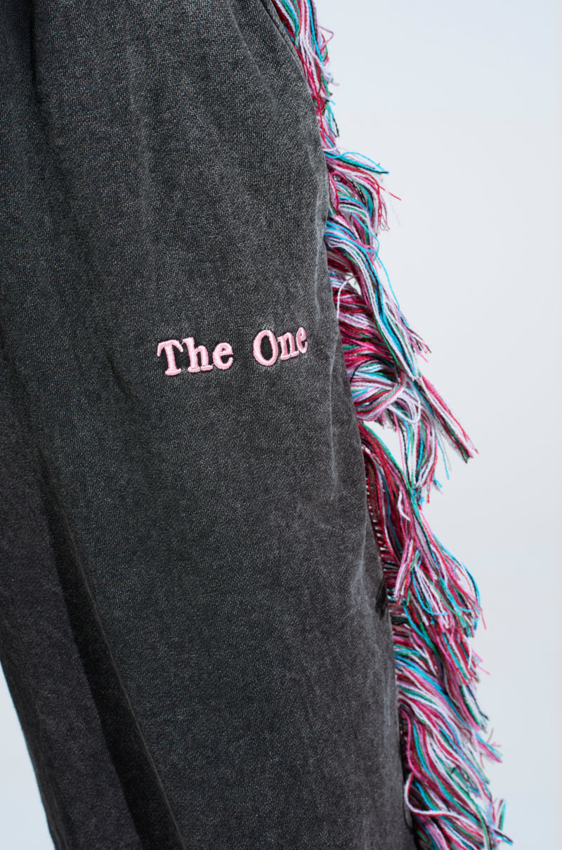 Close-up of a dark fabric with "The One" embroidered in pink, evoking the iconic THE ONE AND ONLY JOGGER. Colorful fringe detailing in blue, pink, and white drapes from the edge on a soft gradient background.