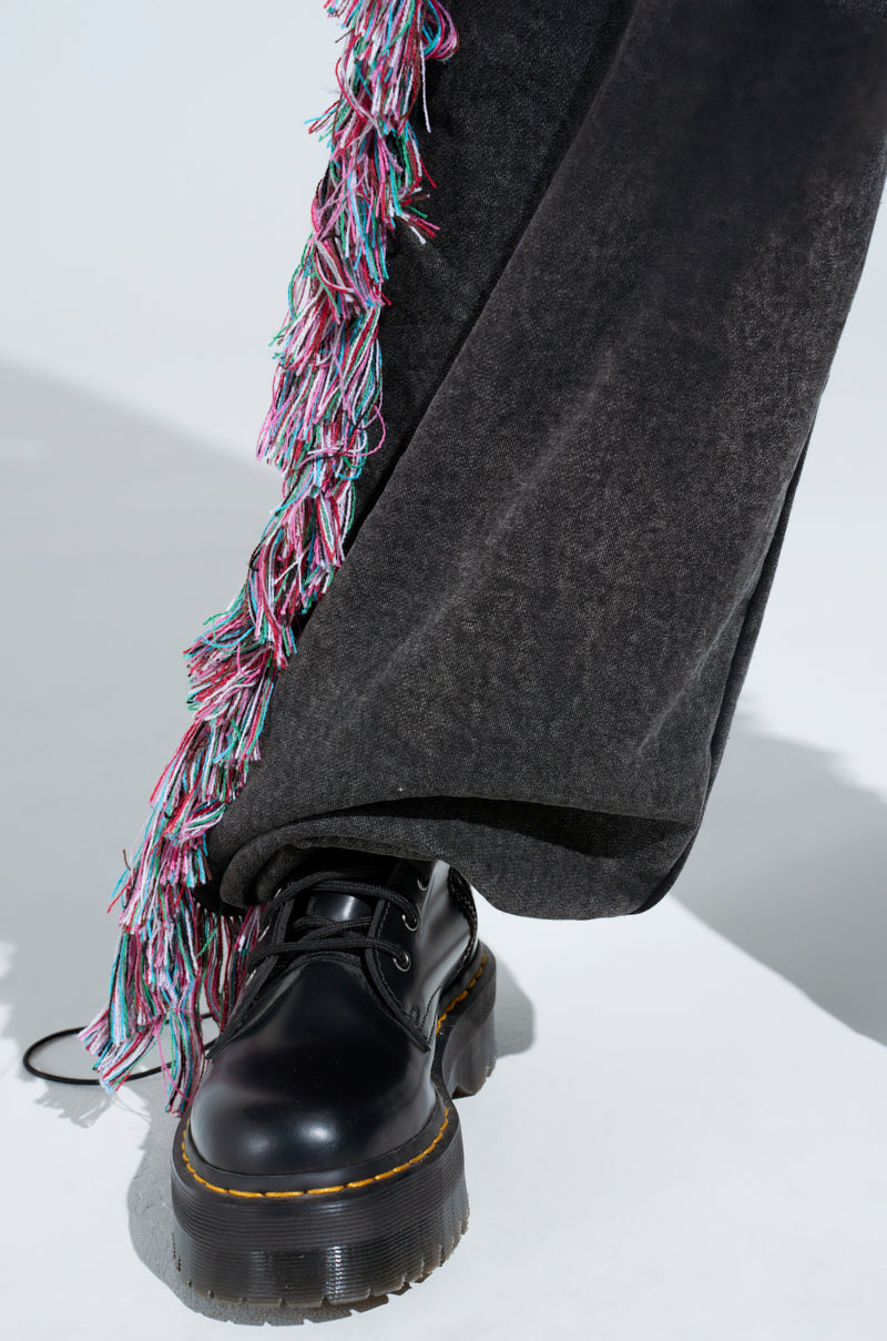A close-up showcases someone wearing THE ONE AND ONLY JOGGER in dark gray, featuring colorful fringe details. They step forward in black platform shoes with thick, textured soles, casting a light shadow on a white surface.
