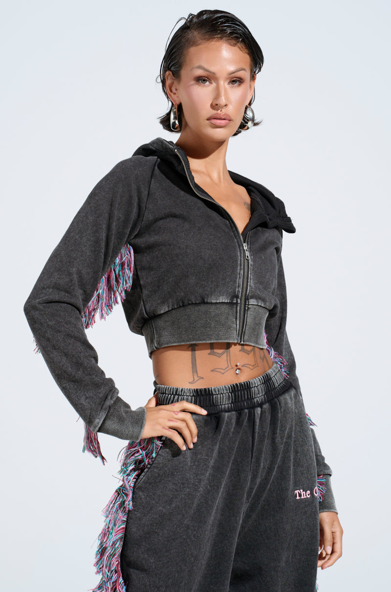 Wearing THE ONE AND ONLY ZIP UP SWEATSHIRT, a person in a dark cropped hoodie with multicolored fringe sleeves and matching sweatpants poses confidently against a light background. Their short, wet-look hair and hoop earrings enhance their self-assured expression.