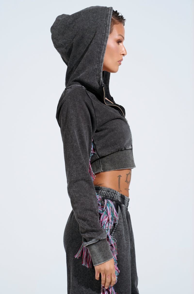 In side profile, a person wears an AKIRA Label dark gray cotton-blend "THE ONE AND ONLY ZIP UP SWEATSHIRT" with the hood raised and matching joggers. Colorful fringe accents enhance the look, revealing short hair and a torso tattoo against a plain light background.