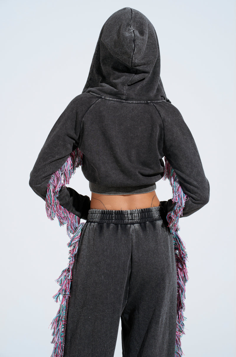 The person, back to the camera, wears the AKIRA Label's bold THE ONE AND ONLY ZIP UP SWEATSHIRT. Its cropped silhouette and dark hood feature colorful fringes on sleeves and sides, all set against a plain light background that highlights the standout design.