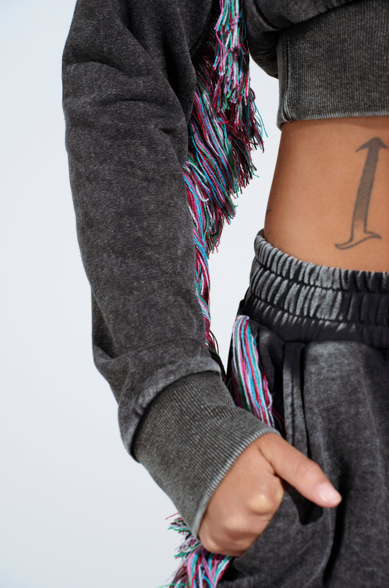 A person wearing THE ONE AND ONLY ZIP UP SWEATSHIRT from AKIRA Label is depicted in a gray cotton-blend with multicolored fringe, shown torso down. Their hand in a pocket reveals a side tattoo against the plain background that highlights the chic outfit and detailed tattoo design.