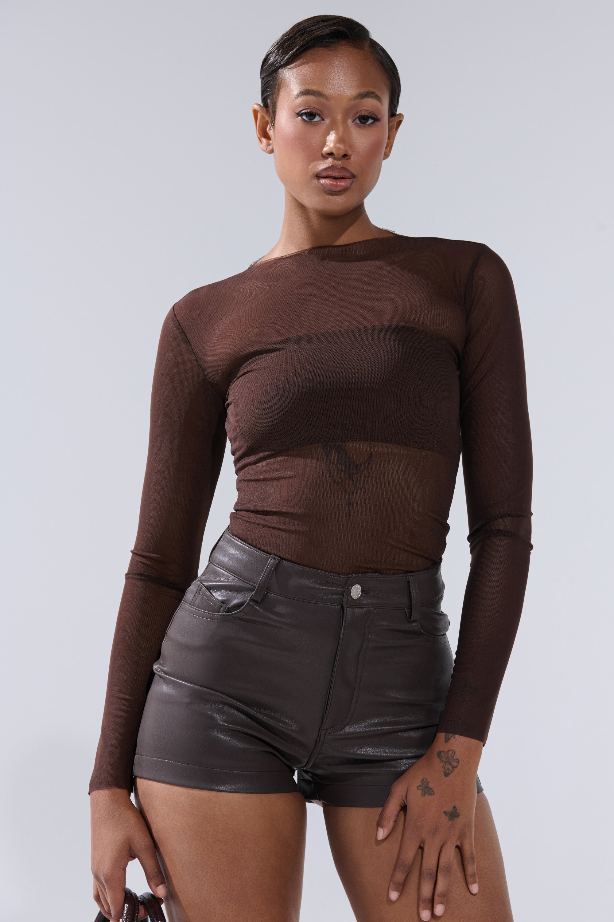 A woman with short hair and tattoos on her left hand wears the PAXTON RAW HEM LONG SLEEVE MESH TEE IN CHOCOLATE BROWN, styled with high-waisted dark leather shorts, standing confidently against a light gray background.