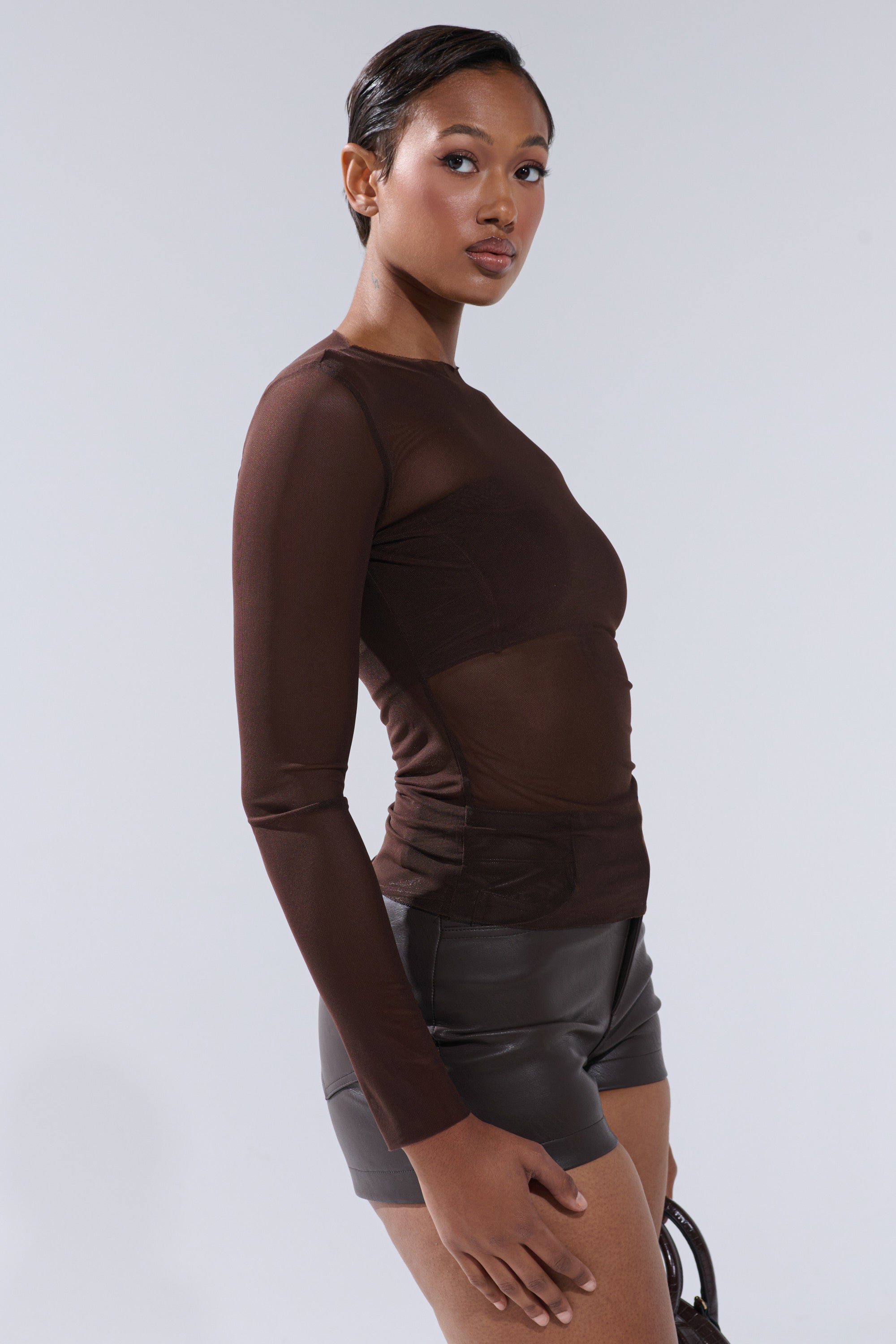 A woman with short hair models the Paxton Raw Hem Long Sleeve Mesh Tee in Chocolate Brown, paired with black leather shorts, standing sideways and looking at the camera against a plain light background.
