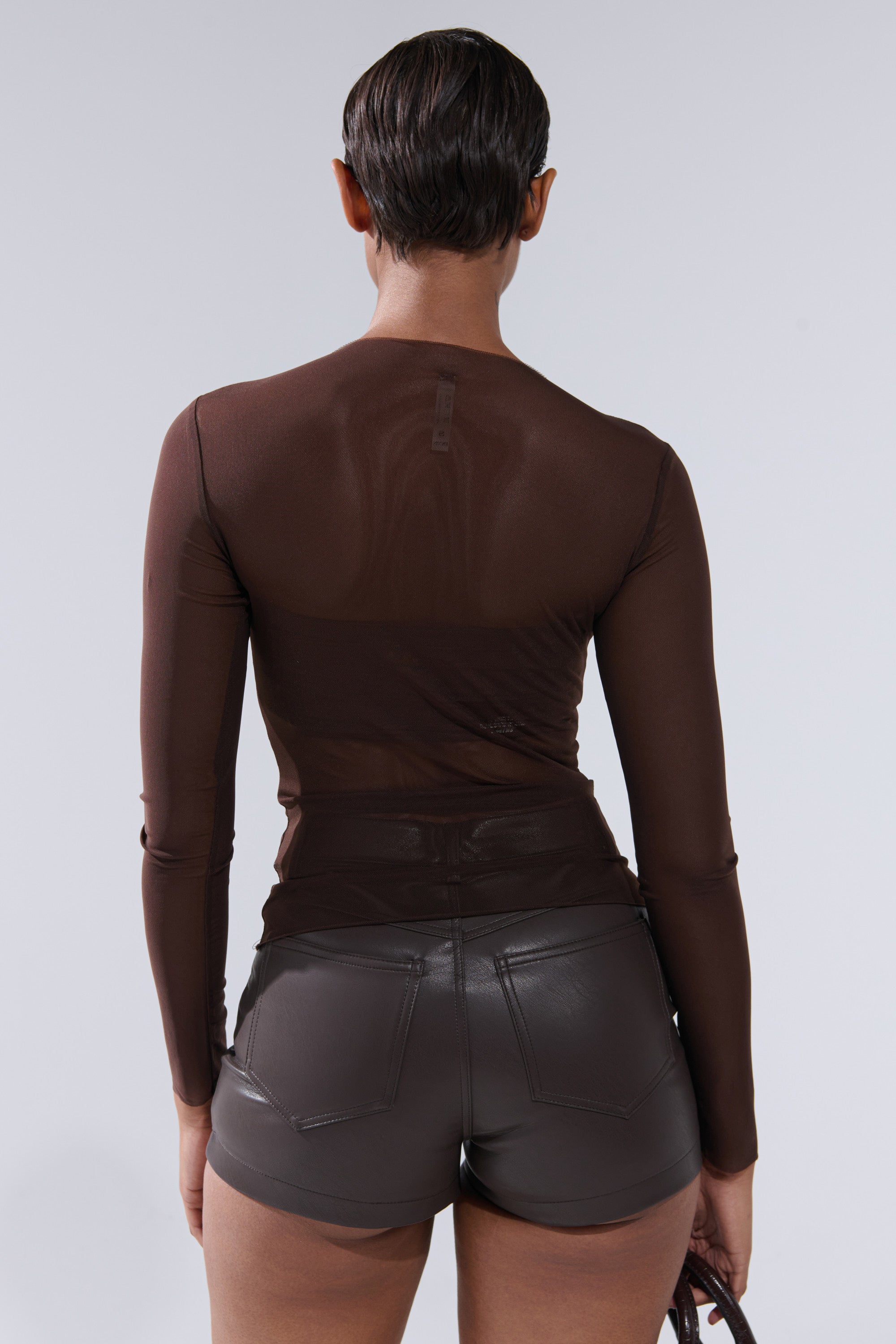 A person with short, slicked-back hair is seen from behind wearing the PAXTON RAW HEM LONG SLEEVE MESH TEE IN CHOCOLATE BROWN and high-waisted, dark leather shorts, standing against a plain light gray background.