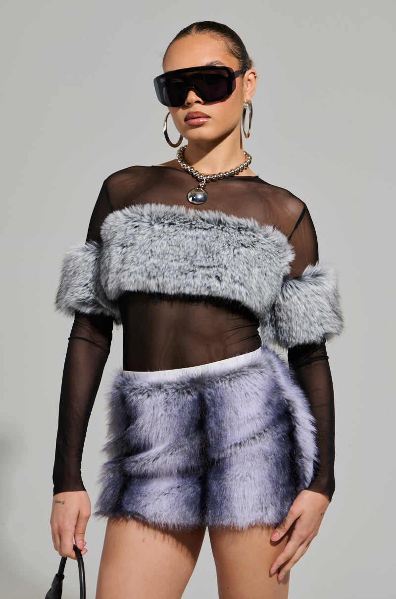 A person gracefully showcases a sheer black JENNY LONG SLEEVE TOP WITH FUR, complemented by a matching mini skirt. They accessorize with bold sunglasses, hoop earrings, and a pearl pendant for added style. Confidently carrying a black purse, they pose against the gray backdrop.