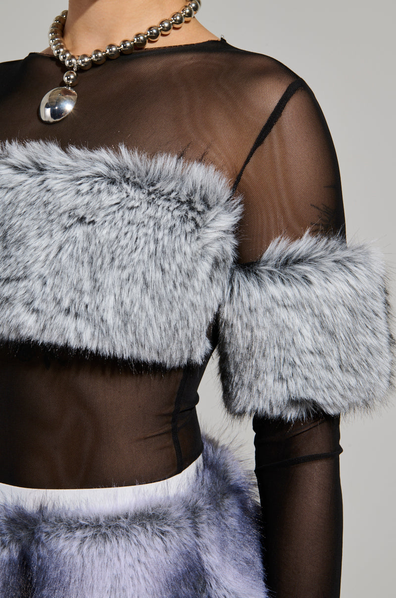 A person wearing the JENNY LONG SLEEVE TOP WITH FUR, featuring a sheer black design with gray faux fur accents on the chest and arms, is paired with a necklace of large, round silver beads. The neutral gray background complements the stylish ensemble.
