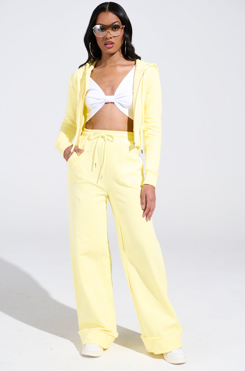 A person in a yellow AKIRA Label BUTTERFINGER SWEATSHIRT with wide-leg pants and a white bikini top. They have long dark hair, sunglasses, and white sneakers, presenting a striking monochromatic look on a light background.