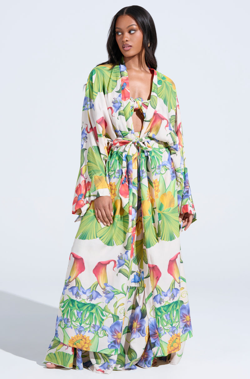 A woman with long hair poses confidently against a plain light background, wearing the INTO THE GARDEN CARDIGAN—a long, flowing open-front piece with a vibrant tropical floral print in green, yellow, blue, red, and purple.