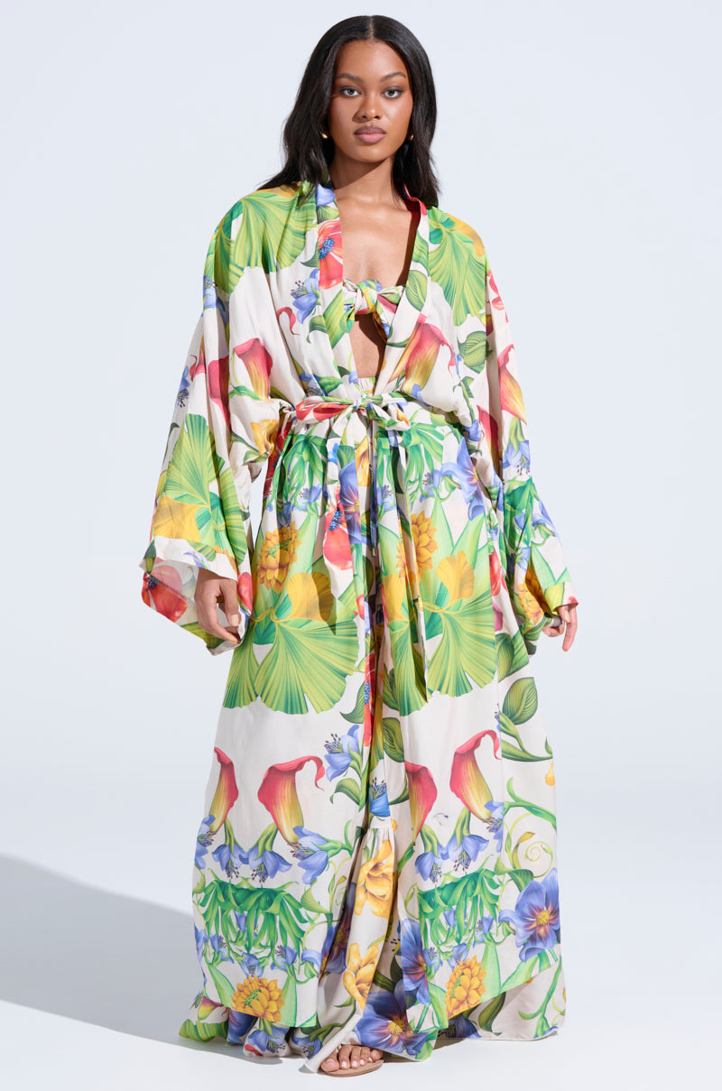 A woman with long dark hair wears the INTO THE GARDEN CARDIGAN over a vibrant, tropical floral dress with wide sleeves. She gazes forward with a neutral expression, completing her breezy, colorful look.