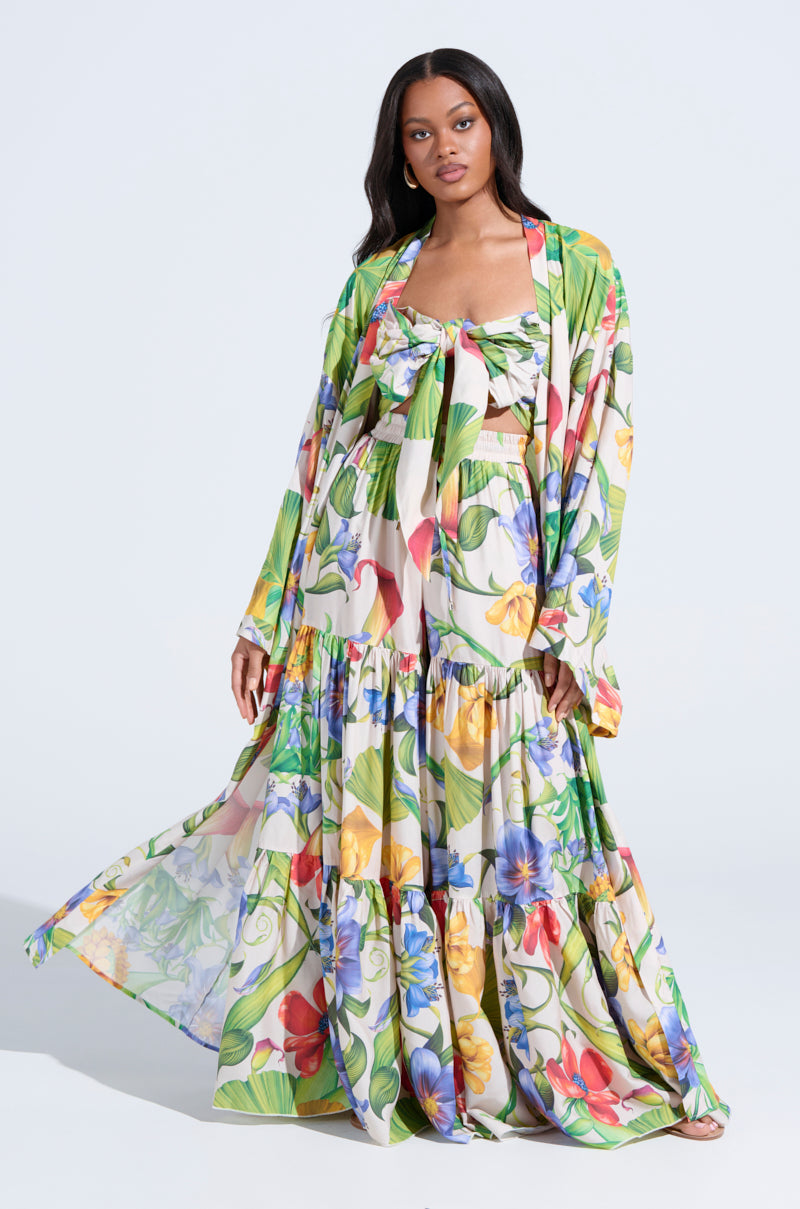 Wearing the INTO THE GARDEN CARDIGAN over a flowing, ankle-length tropical floral print skirt and matching bow-front crop top, a woman stands confidently against a light background.