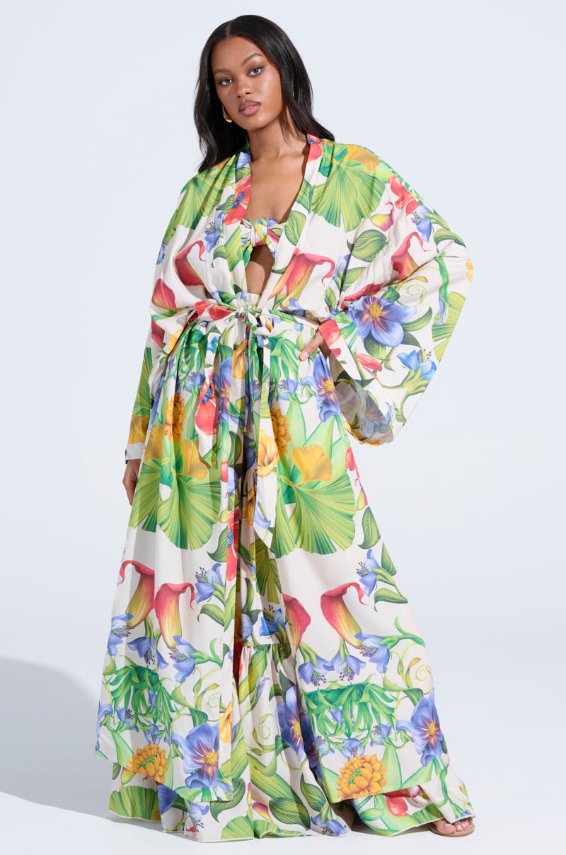A woman models the INTO THE GARDEN CARDIGAN, a long, flowing open-front piece with wide sleeves and a vibrant tropical floral print in green, yellow, red, and blue, standing against a plain light background.