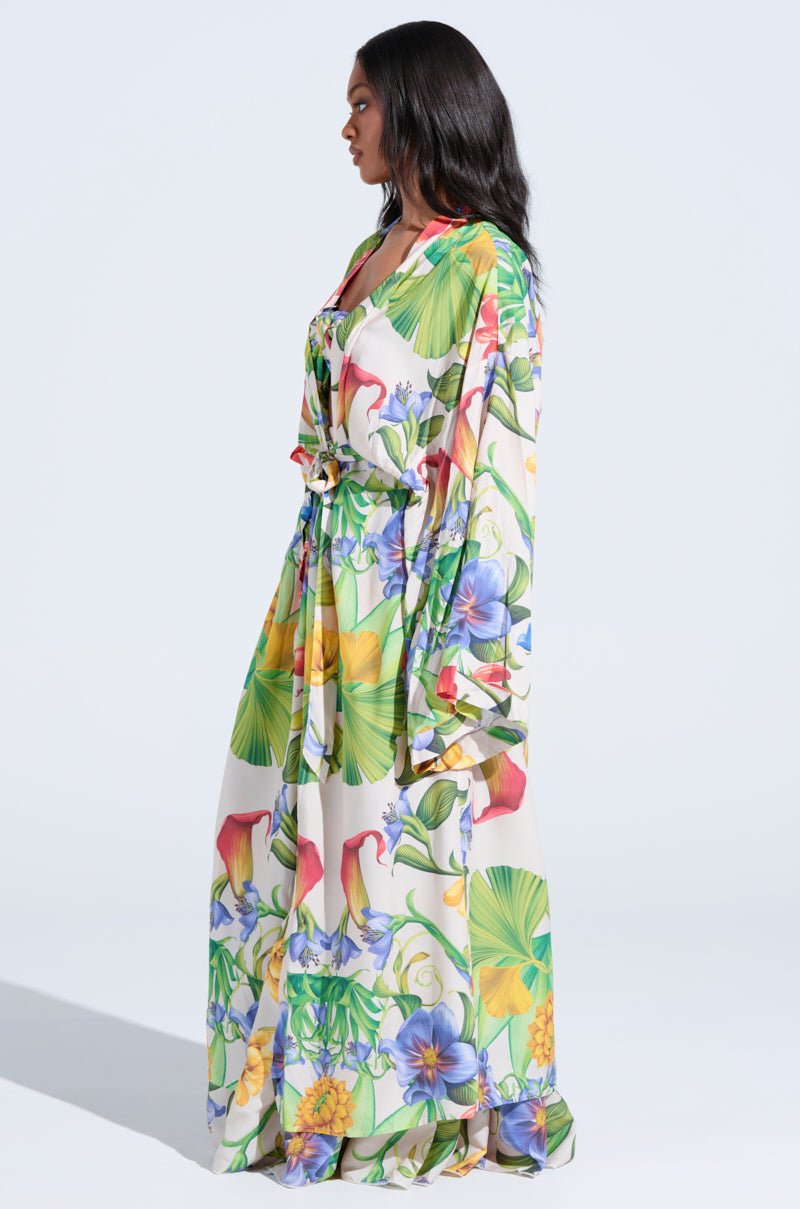 A woman stands in profile wearing the INTO THE GARDEN CARDIGAN over a long, tropical floral dress with green leaves and vibrant flowers. The background is plain light blue.
