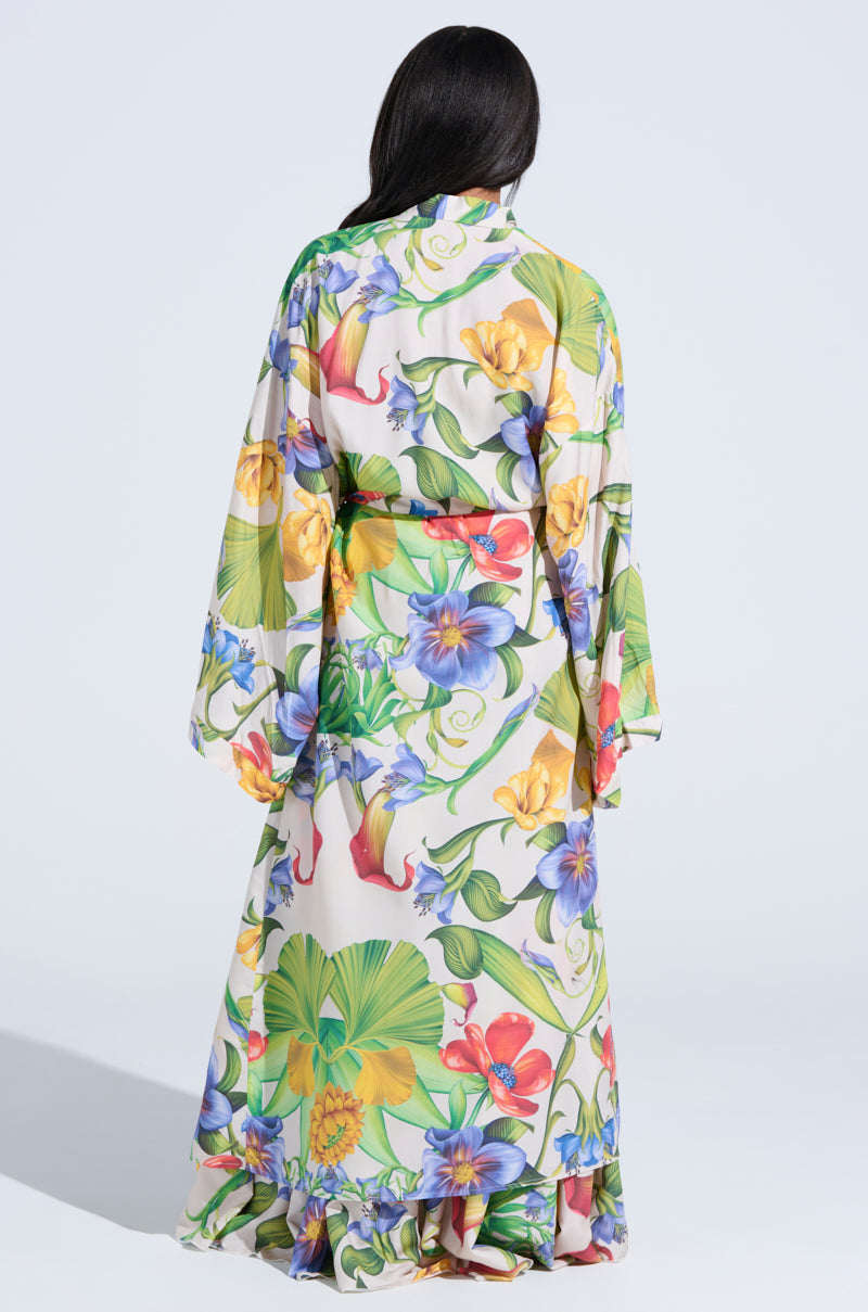 A woman with long dark hair faces away, wearing the INTO THE GARDEN CARDIGAN – a lightweight piece with wide sleeves and a vibrant tropical floral print on a light background.