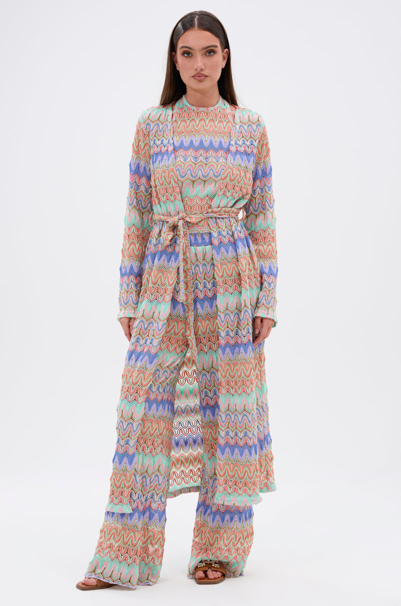 A woman wears the TAKE ME TO MIAMI DUSTER—a colorful, long-sleeved, zigzag-patterned belted maxi dress with matching wide-leg pants and brown sandals—posing against a plain white background.