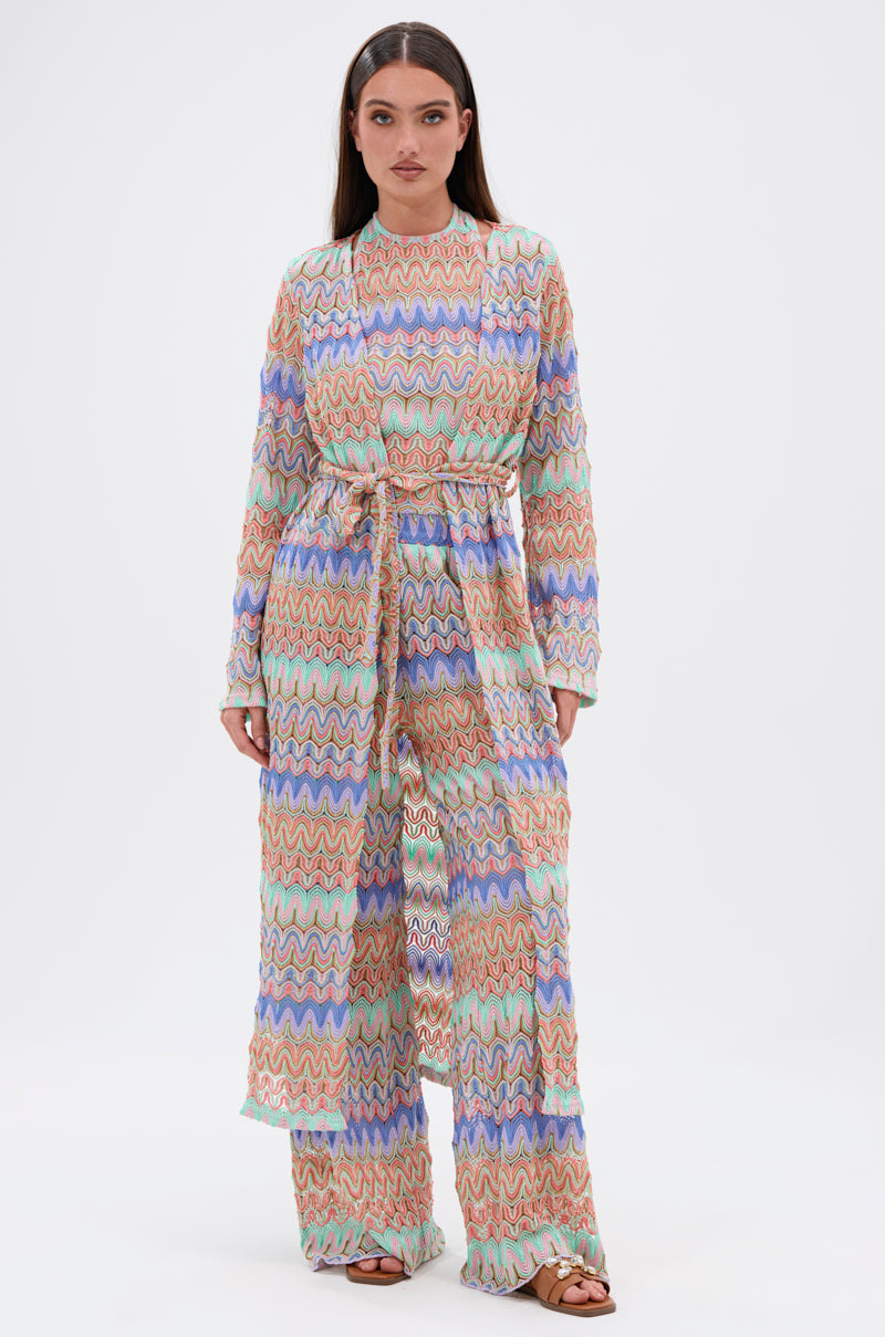 A woman faces forward in the TAKE ME TO MIAMI DUSTER, featuring wavy colorful patterns and a belted waist, paired with matching wide-leg pants. She styles the look with long straight hair and brown sandals against a white background.
