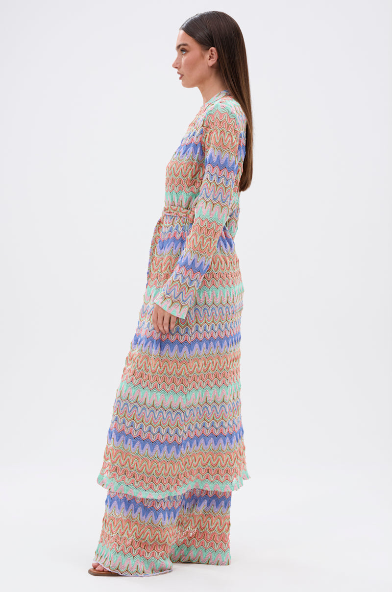 A woman stands in profile wearing the TAKE ME TO MIAMI DUSTER, a long, colorful crochet dress with a wavy zigzag pattern in pastel orange, blue, purple, and green. The resort-ready look features long sleeves and a tiered flowing skirt.
