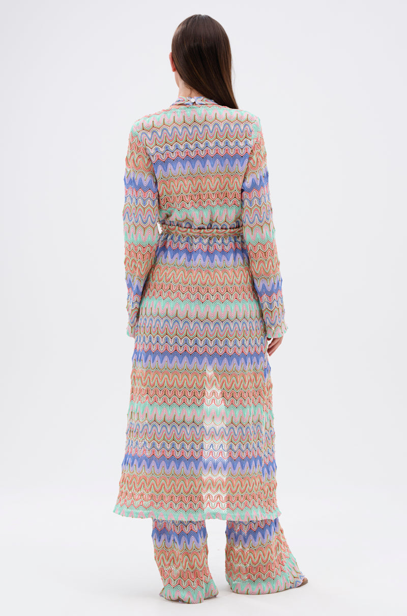 A woman stands facing away, wearing the TAKE ME TO MIAMI DUSTER and matching wide-leg pants with vibrant wavy blue, green, pink, and red patterns on white—her straight brown hair flowing loosely for a perfect resort-ready look.