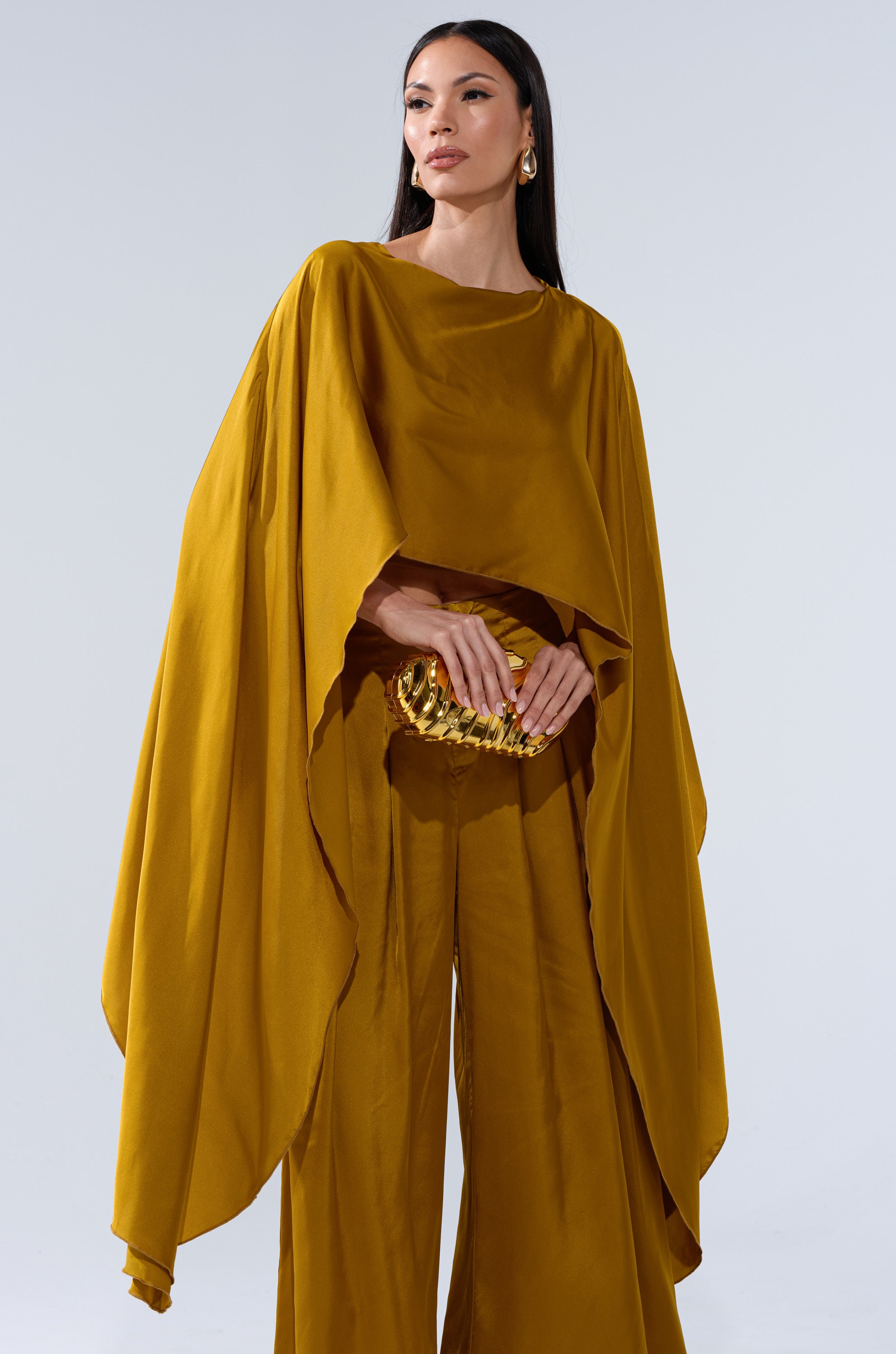 A woman in the COCKTAIL HOUR BLOUSE IN MUSTARD, a flowing satin top with wide sleeves and a high-low hem, paired with matching wide-leg pants and a gold clutch, stands against a plain light gray background.