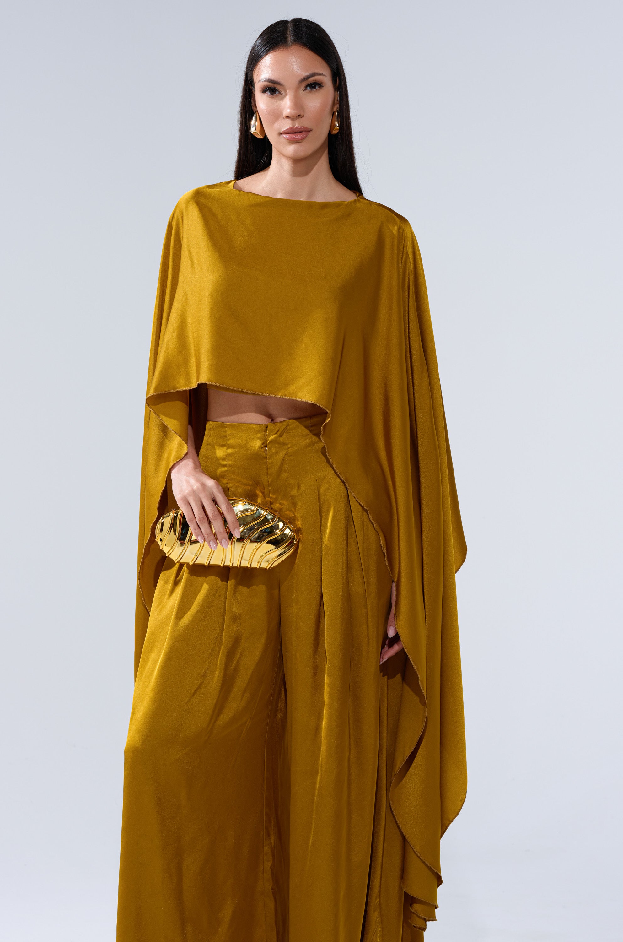 A woman with long, straight dark hair and gold earrings poses against a plain light background, wearing the COCKTAIL HOUR BLOUSE IN MUSTARD with wide-legged pants and holding a shiny gold clutch.