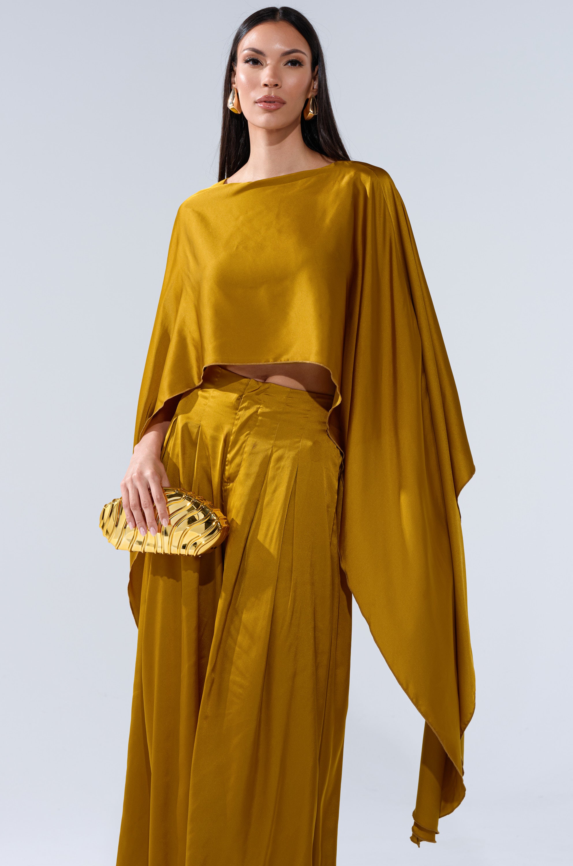 A woman in a flowing COCKTAIL HOUR BLOUSE IN MUSTARD with a high-low hem and matching wide-leg pants holds a gold clutch. She has long, straight dark hair, gold earrings, and stands against a plain light background.