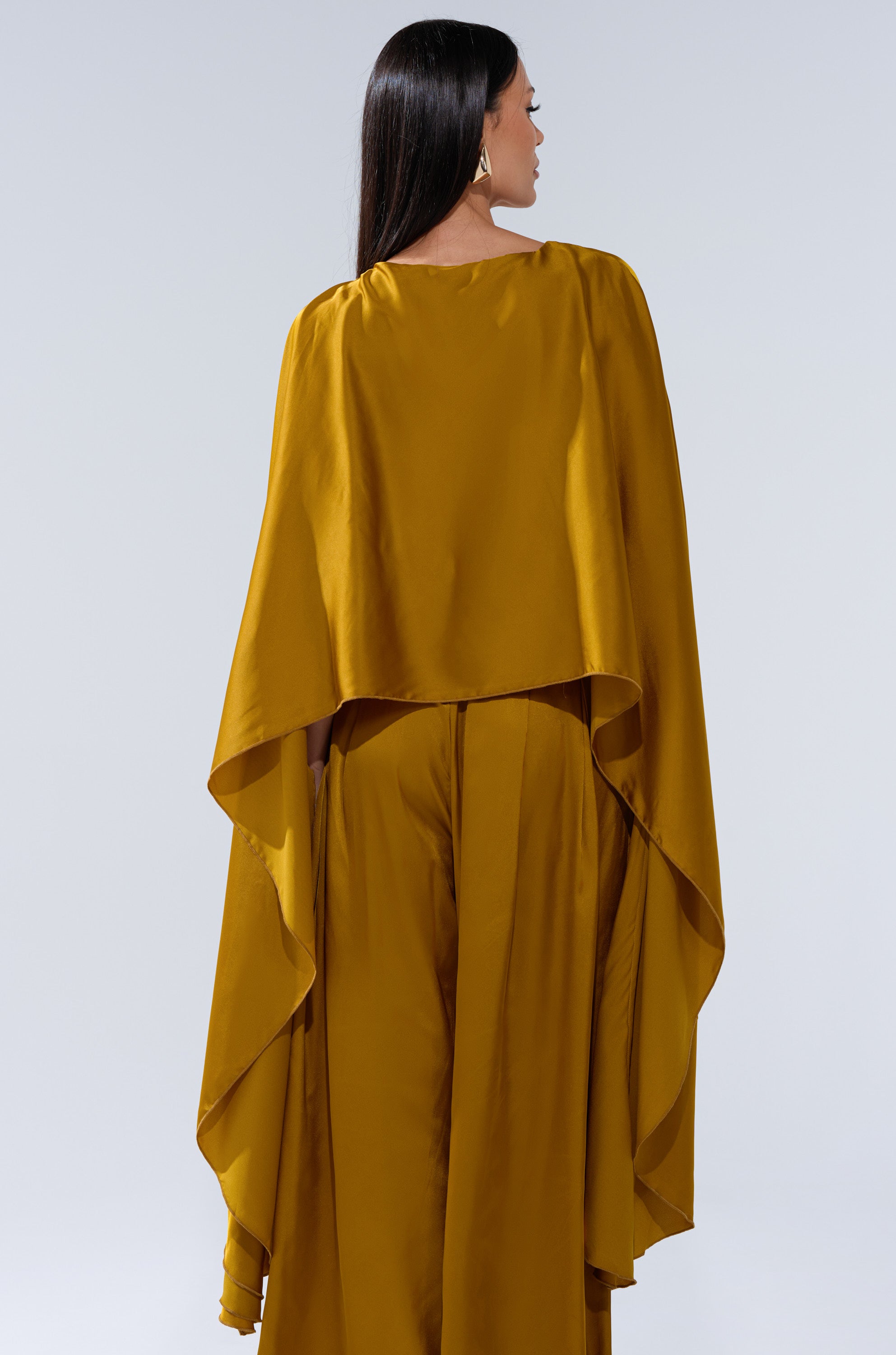 A woman with long dark hair, seen from behind, wears the COCKTAIL HOUR BLOUSE IN MUSTARD— a loose-fitting satin blouse with cape-like sleeves and a high-low hem—paired with wide-leg pants against a light gray background.