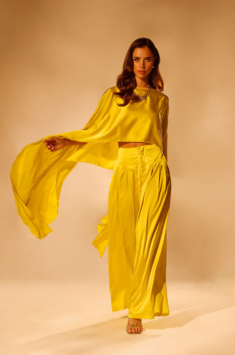 A woman poses in COCKTAIL HOUR WIDE LEG PANT—a bright yellow satin, high-rise, wide-leg style—paired with a draped top and strappy heeled sandals against a warm beige background.