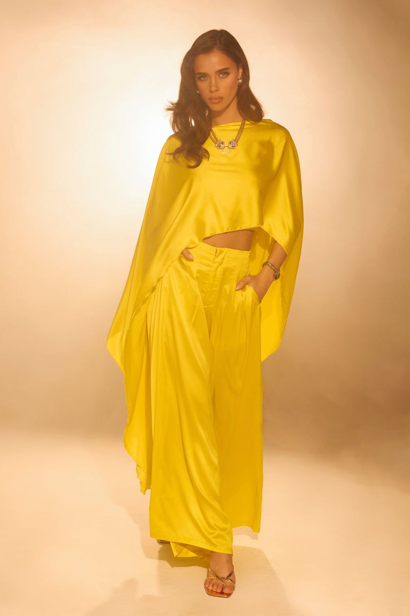 A woman poses confidently in the COCKTAIL HOUR WIDE LEG PANT, a silky satin yellow style with a high-rise waistband and flowing top, accessorized with a statement necklace and heels against a warmly lit background.