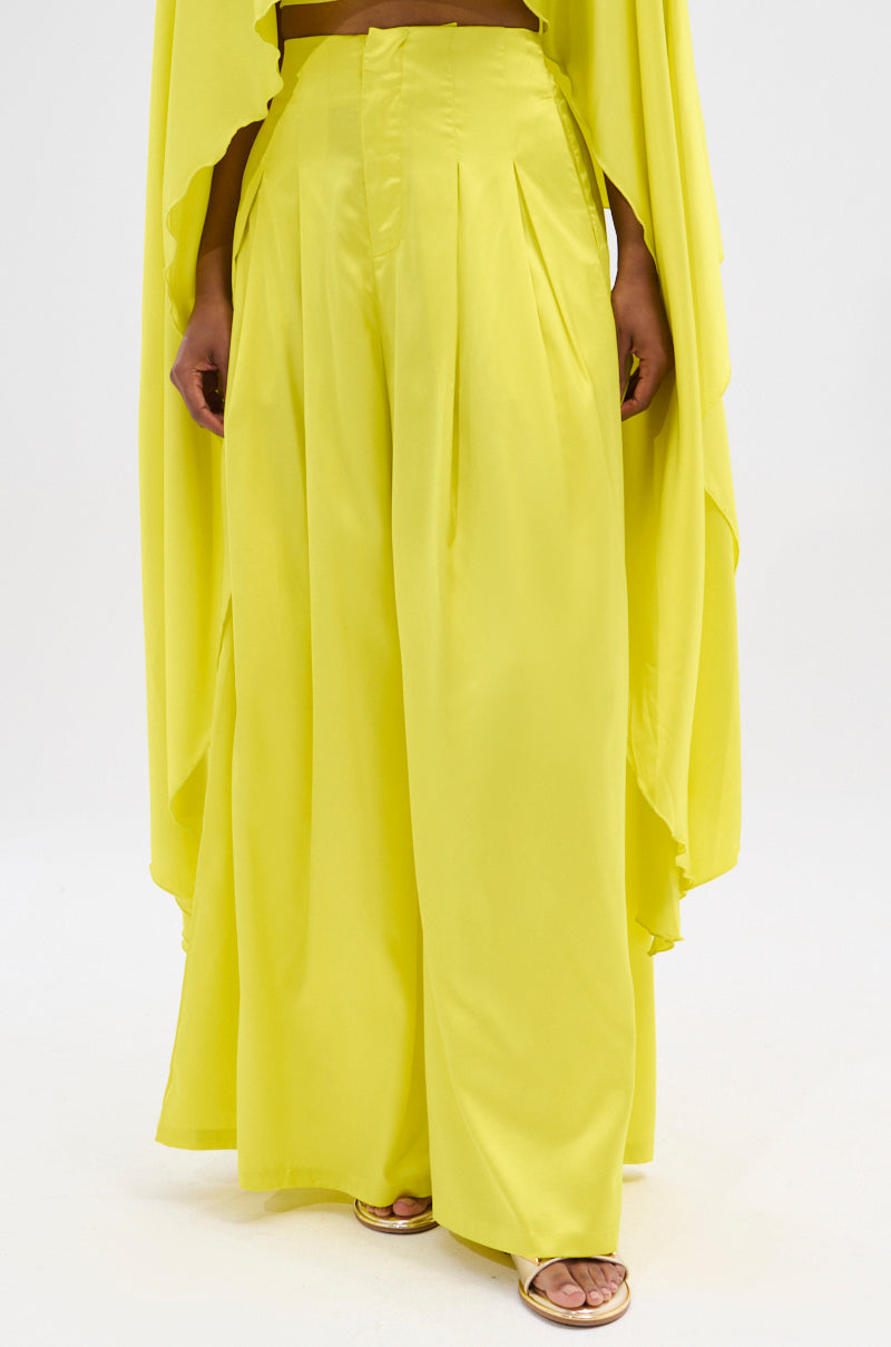 The person confidently dons vibrant wide-legged yellow pants and a matching flowy top, echoing the COCKTAIL HOUR WIDE LEG PANT style. The satin-like fabric drapes elegantly with a trailing hem, completing the chic look when paired with stylish sandals.