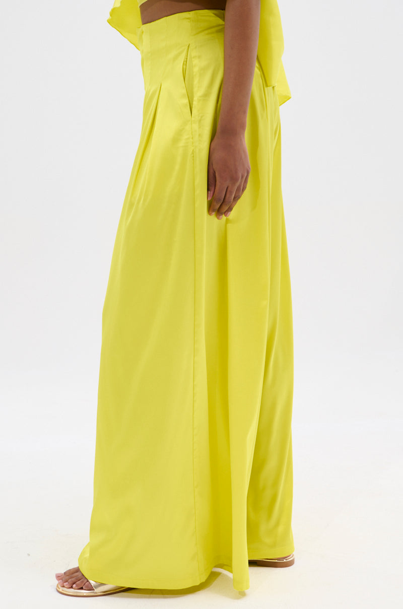 A person stands sideways in bright yellow COCKTAIL HOUR WIDE LEG PANTs and an off-shoulder top, their hand resting on the flowing satin. White sandals complete the look against a plain, light background that highlights their vibrant style.