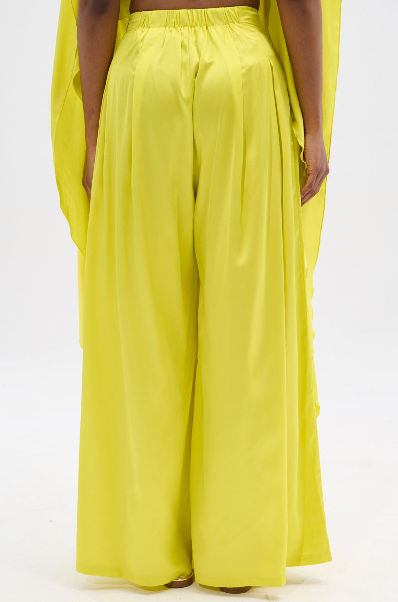 A person wears the COCKTAIL HOUR WIDE LEG PANT in bright yellow, beautifully gathered at the waist. The matching draped fabric over their shoulders completes this chic look against a plain white backdrop.