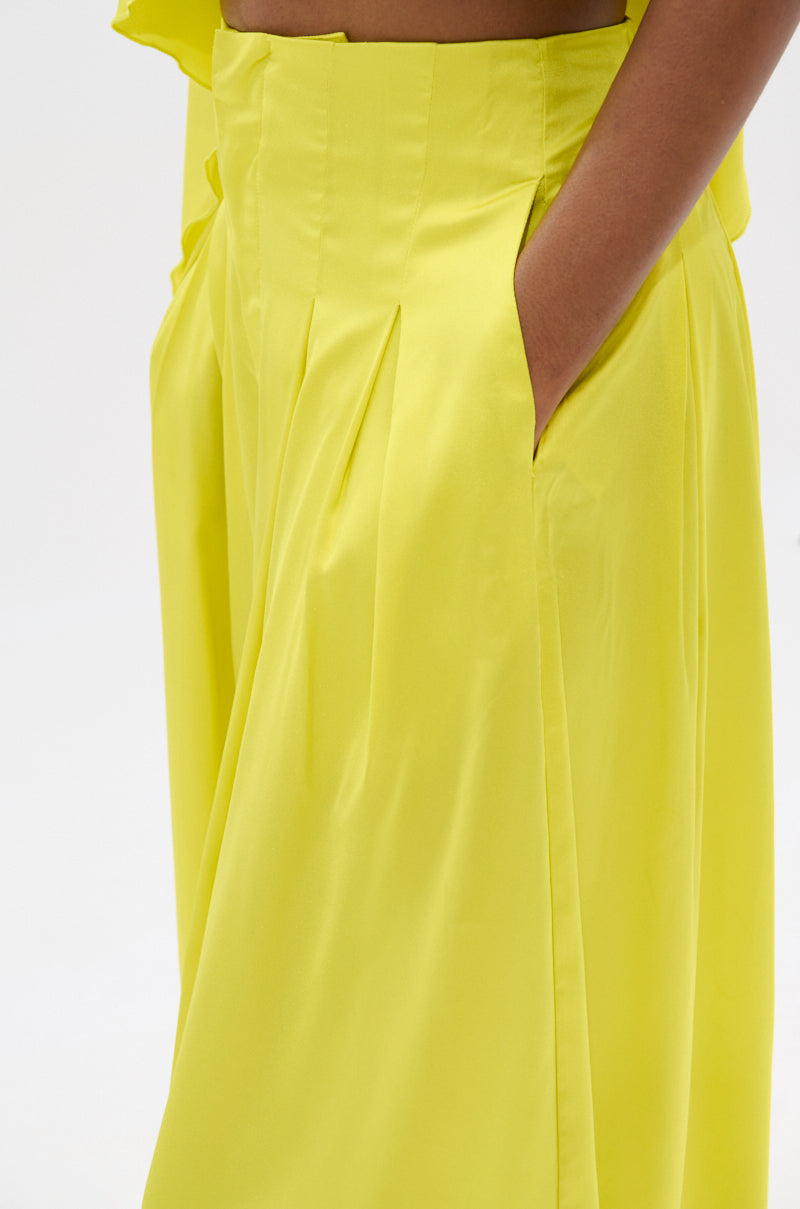 A close-up shows a person wearing bright yellow COCKTAIL HOUR WIDE LEG PANT with their right hand in the pocket, the smooth and shiny fabric contrasting against a plain white background.