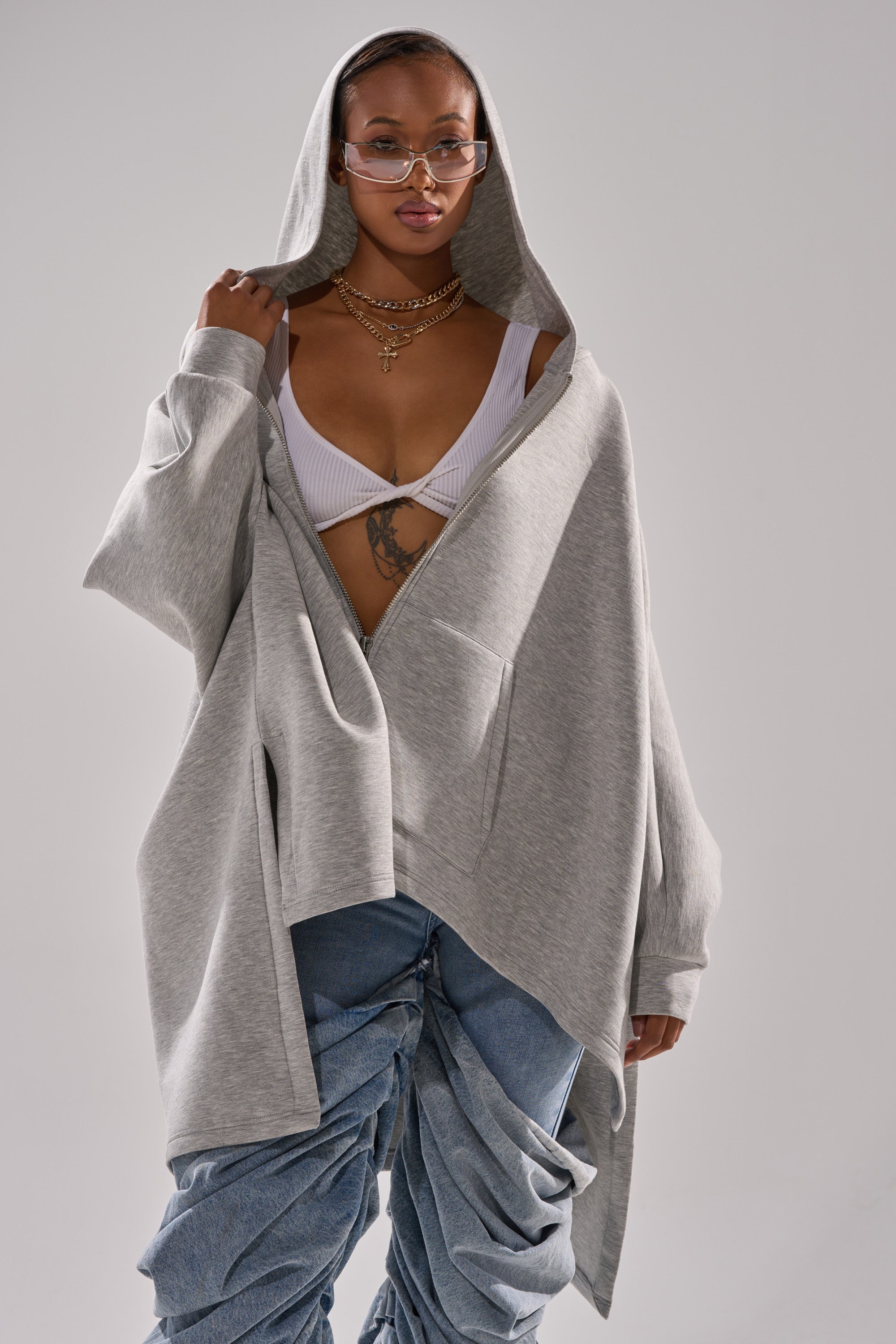 A woman wearing glasses, layered necklaces, a white bralette, the CREW LOVER OVERSIZED ZIP UP SWEATSHIRT in heather grey, and ruched denim pants stands facing forward, holding her hood with one hand.